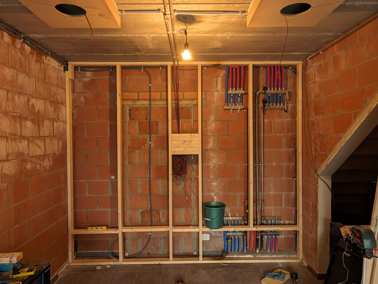 A wooden stud wall in front of a brick wall with a water manifold